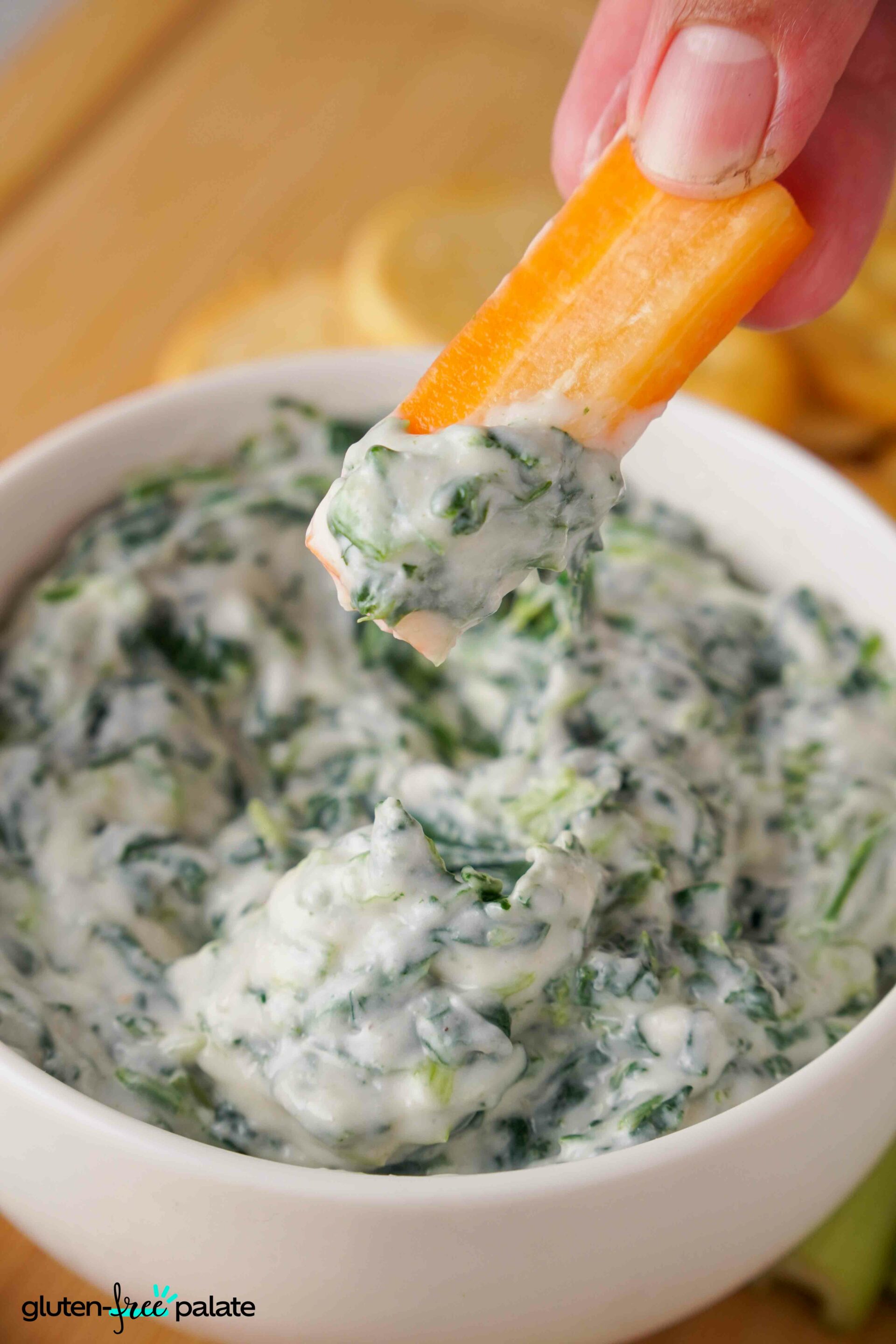 Spinach dip in a white bowl with a carrot being dipped into it.