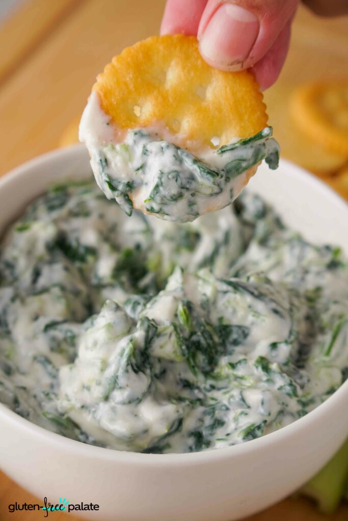 Spinach dip in a white bowl with a cracker being dipped into it.