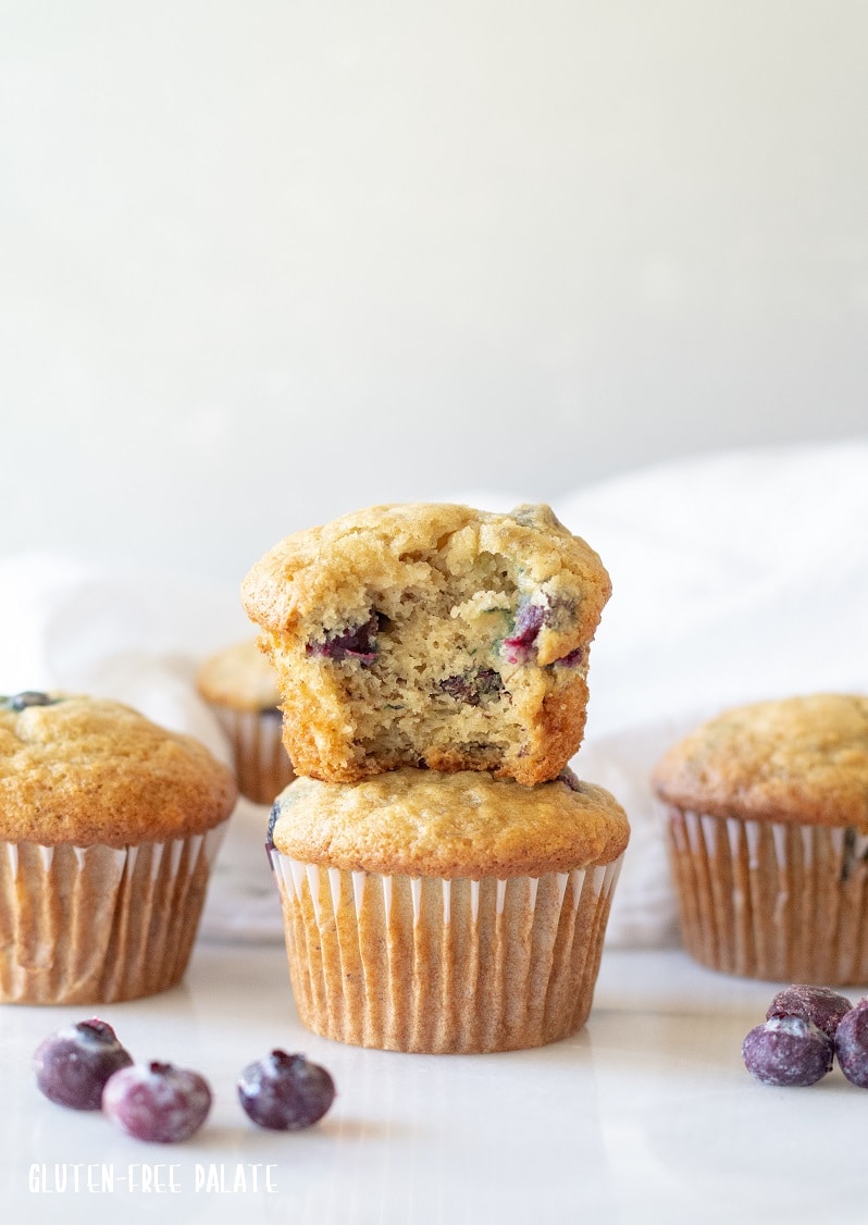 Gluten Free Blueberry Banana Muffins stacked on top of one another, one has a bite out, they are stacked near three other muffins and scattered blueberries