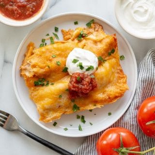 two enchiladas with cheese, sour cream, and salsa on a white plate next to a two tomatoes and a bowl of salsa and sour cream