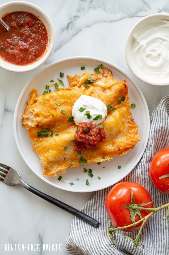 two enchiladas with cheese, sour cream, and salsa on a white plate next to a two tomatoes and a bowl of salsa and sour cream
