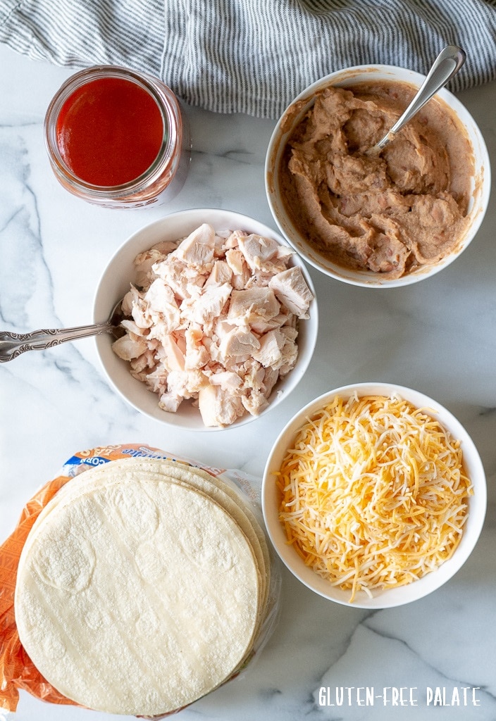 a stack of corn tortillas, a bowl of shredded chicken, a bowl of shredded cheese, a bowl of refried beans, a jar of enchilada sauce