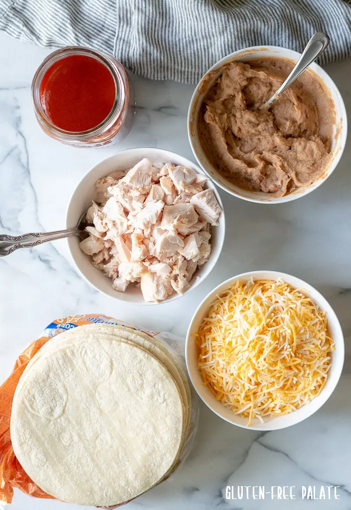 a stack of corn tortillas, a bowl of shredded chicken, a bowl of shredded cheese, a bowl of refried beans, a jar of enchilada sauce