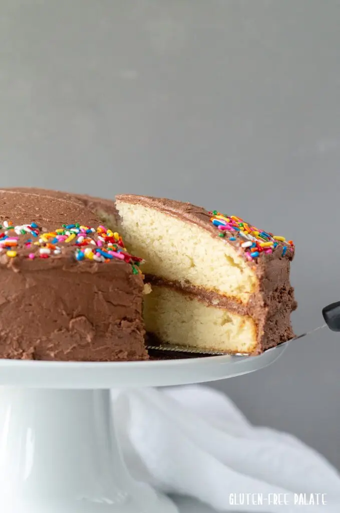 A slice of gluten-free vanilla cake with chocolate frosting being served from a cake plate.