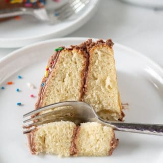 A slice of gluten-free vanilla cake topped with chocolate frosting on a white plate with a fork and colored sprinkles
