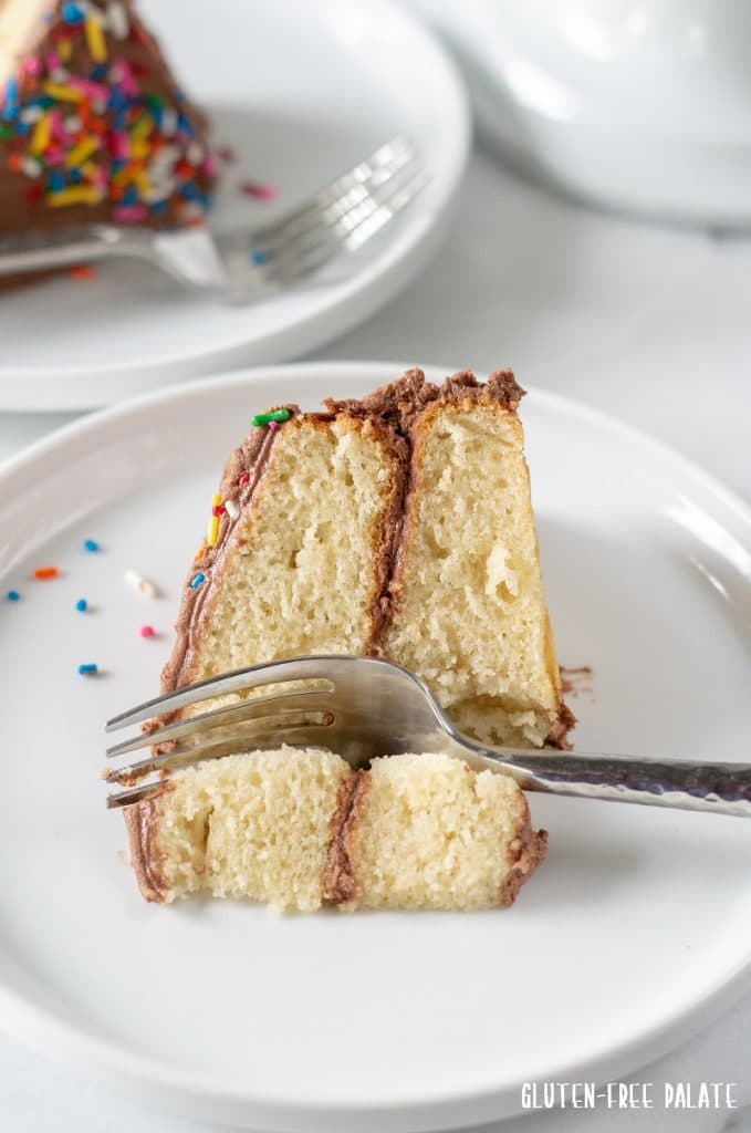 A slice of gluten-free vanilla cake topped with chocolate frosting on a white plate with a fork and colored sprinkles