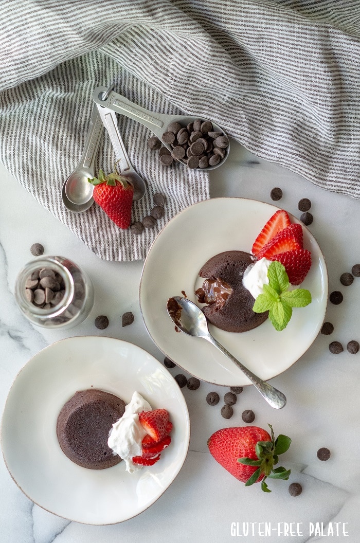 two lava cakes with a chocolate centers topped with whipped cream and strawberries, on white plates with chocolate chips around them