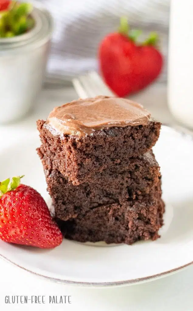 three gluten-free brownies stacked on a white plate with two strawberries