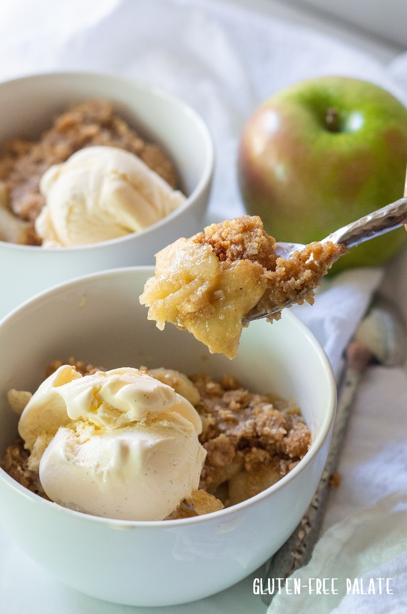 Gluten free apple crisp in a white bowl with ice cream, a spoon is taking a bite out