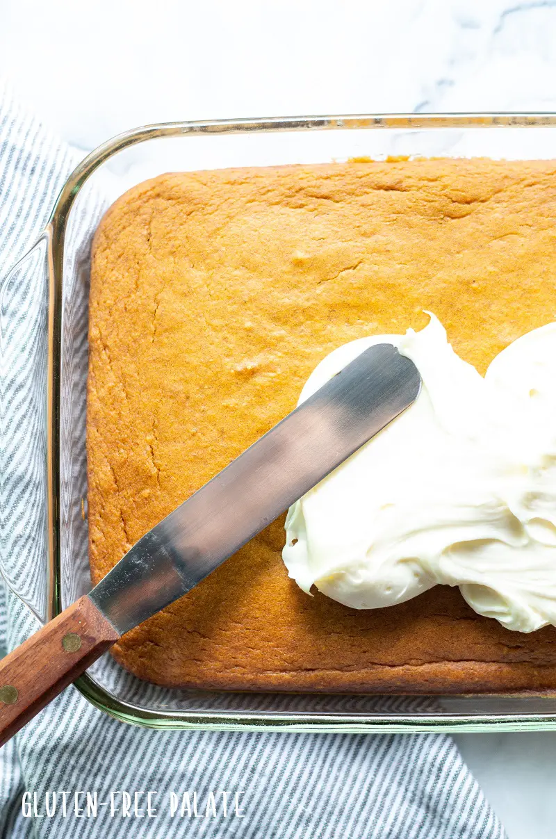 a knife spreading white frosting on gluten free pumpkin cake in a glass baking pan