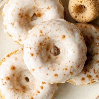 close up of gluten-free apple cider donuts with white icing and cinnamon sprinkled on top on a cream colored plate