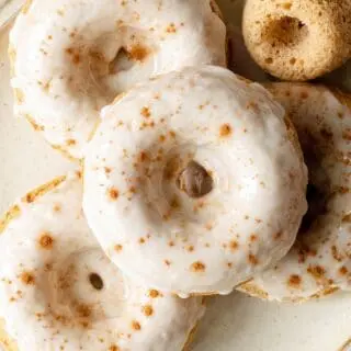 close up of gluten-free apple cider donuts with white icing and cinnamon sprinkled on top on a cream colored plate