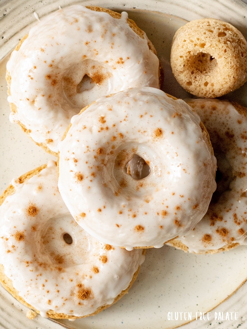 close up of gluten free apple cider donuts with white icing and cinnamon sprinkled on top on a cream colored plate