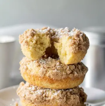 three gluten free crumb donuts stacked on a plate, one has a bite out of it