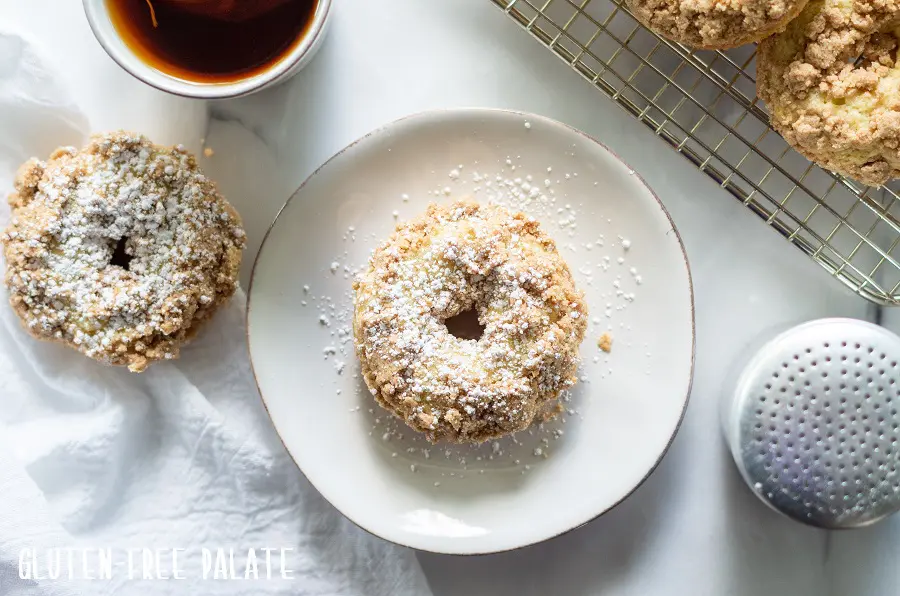 Gluten Free Crumb Donut sprinkled with powdered sugar on a white plate next to a donut, a cup of coffee and a cooling rack