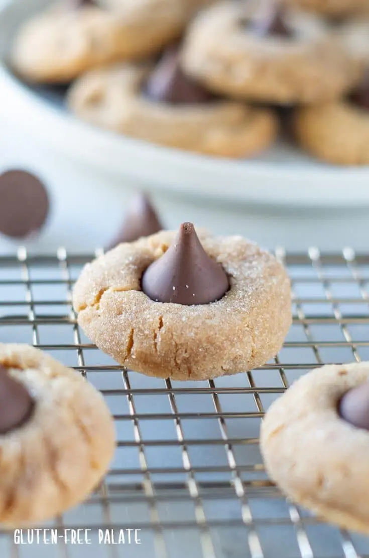 gluten-free peanut butter blossoms with a hershey kiss pressed on top, on a wire rack with a plate of cookies behind it
