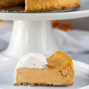 a slice of Gluten-Free Pumpkin Cheesecake on a white plate with pumpkin cheesecake on a white cake stand behind it