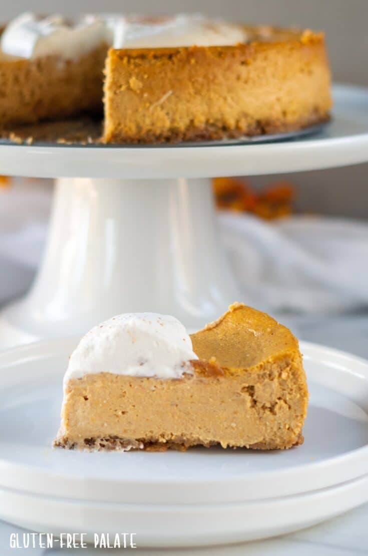a slice of Gluten Free Pumpkin Cheesecake on a white plate with pumpkin cheesecake on a white cake stand behind it
