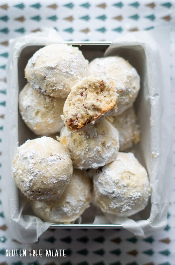 gluten free snow ball cookies in a square metal tin lined with wax paper, one cookie has a bite out