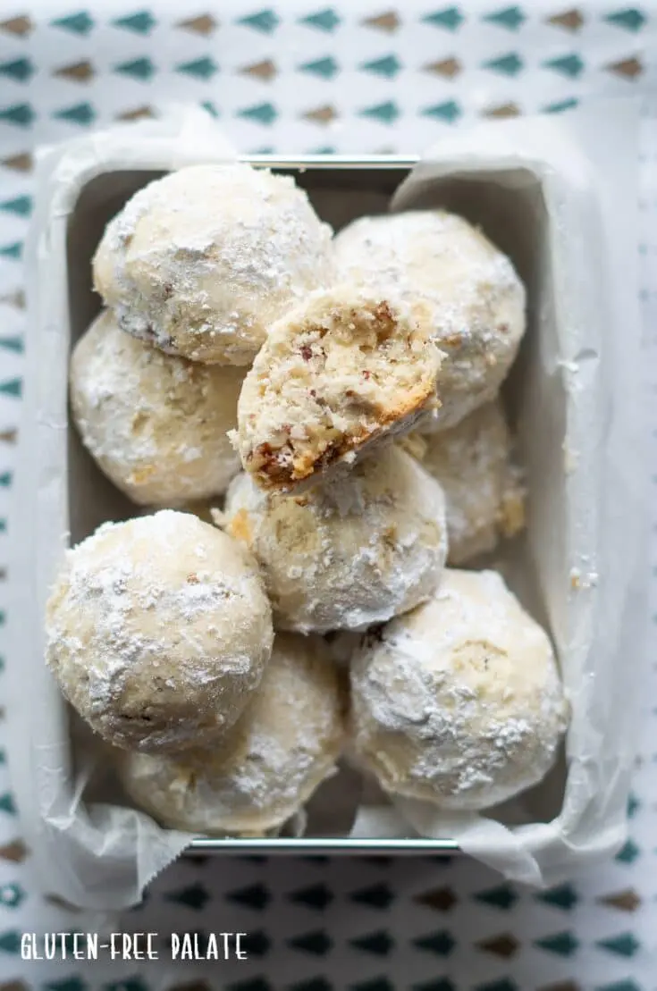 gluten free snow ball cookies in a square metal tin lined with wax paper, one cookie has a bite out