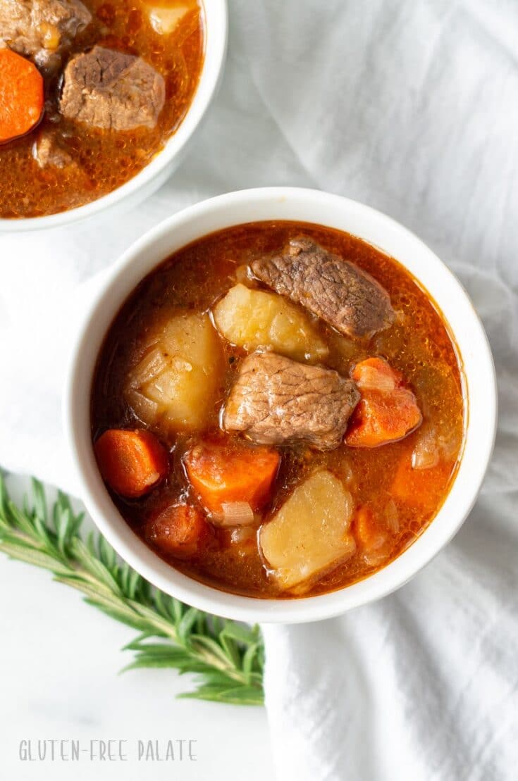 gluten free beef stew in a white bowl with beef, potatoes, and carrots with a spring of rosemary next to it