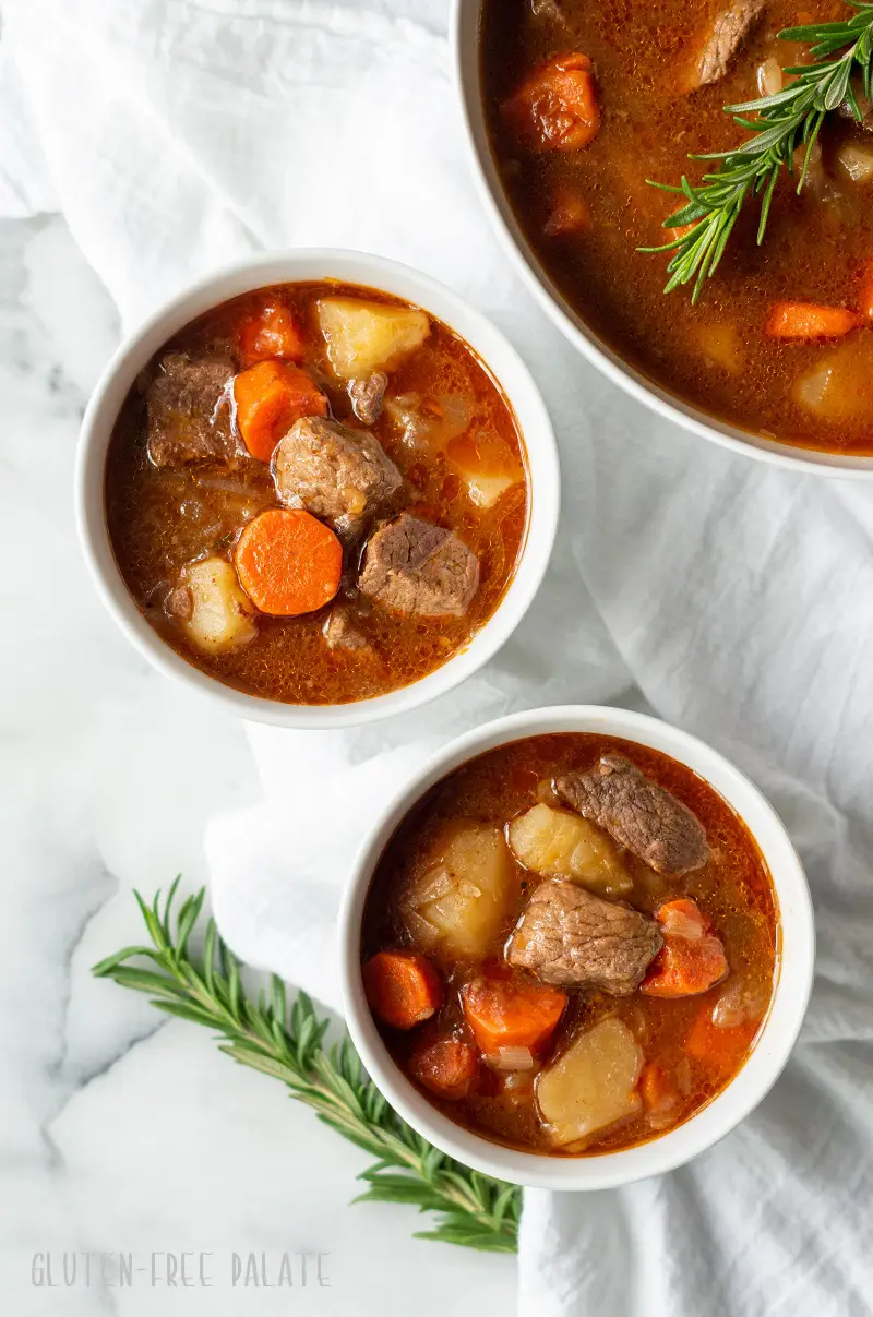 two bowls of gluten-free beef stew next to a pot of beef stew with springs of rosemary next to it.