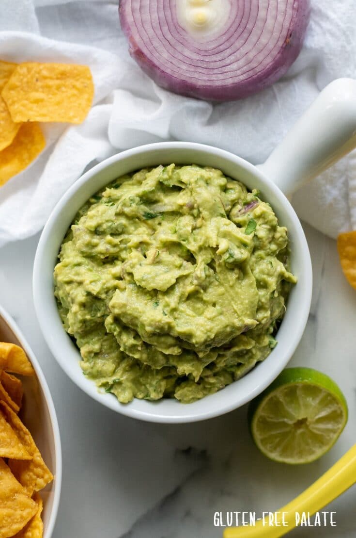 guacamole in a white bowl surrounded by red onion and yellow chips and a lime wedge