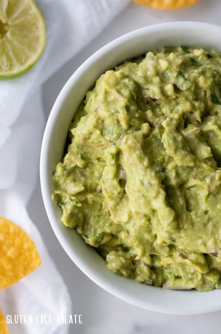 close up of guacamole in a white bowl with a lime wedge and yellow chips