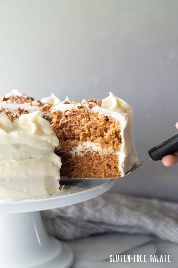 Gluten-free carrot cake with cream cheese frosting being sliced on a white cake stand