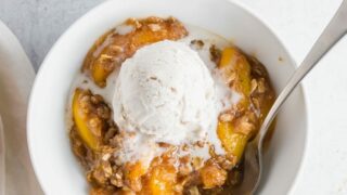 a close up view of a gluten free peach crisp in a white bowl topped with ice cream