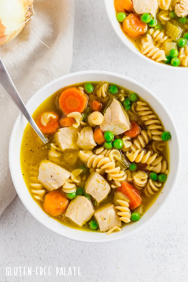 a close up of gluten-free chicken noodle soup in a white bowl with a spoon