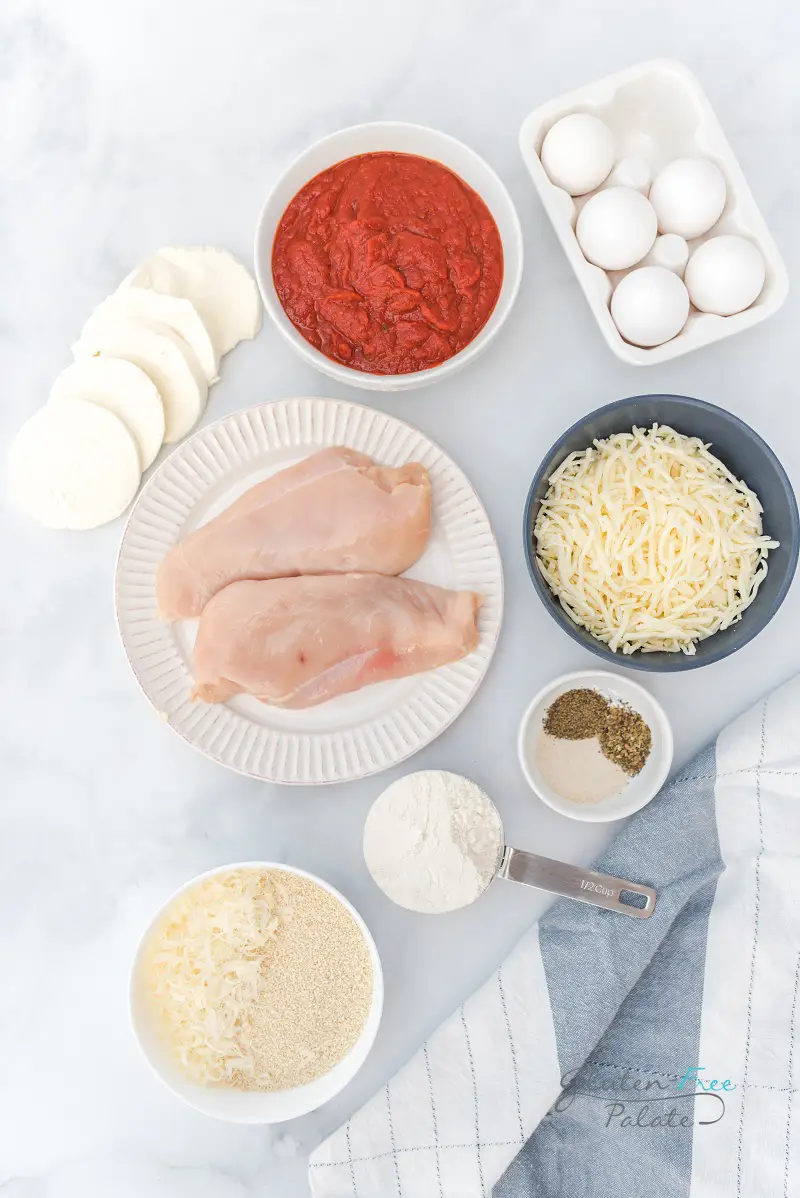 Top down view of the ingredients needed for gluten free chicken parmesan, including chicken, bread crumbs, cheese, sauce, eggs, and seasonings