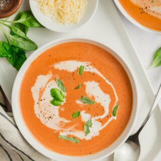 top down view of a bowl of creamy gluten-free tomato soup with a spoon, parmesan cheese, and pepper on the side and garnished with basil.