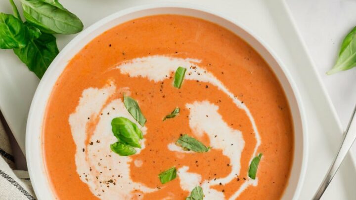 top down view of a bowl of creamy tomato soup with a spoon, parmesan cheese, and pepper on the side and garnished with basil.