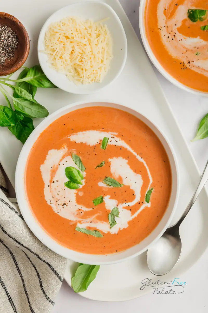 top down view of a bowl of creamy tomato soup with a spoon, parmesan cheese, and pepper on the side and garnished with basil.