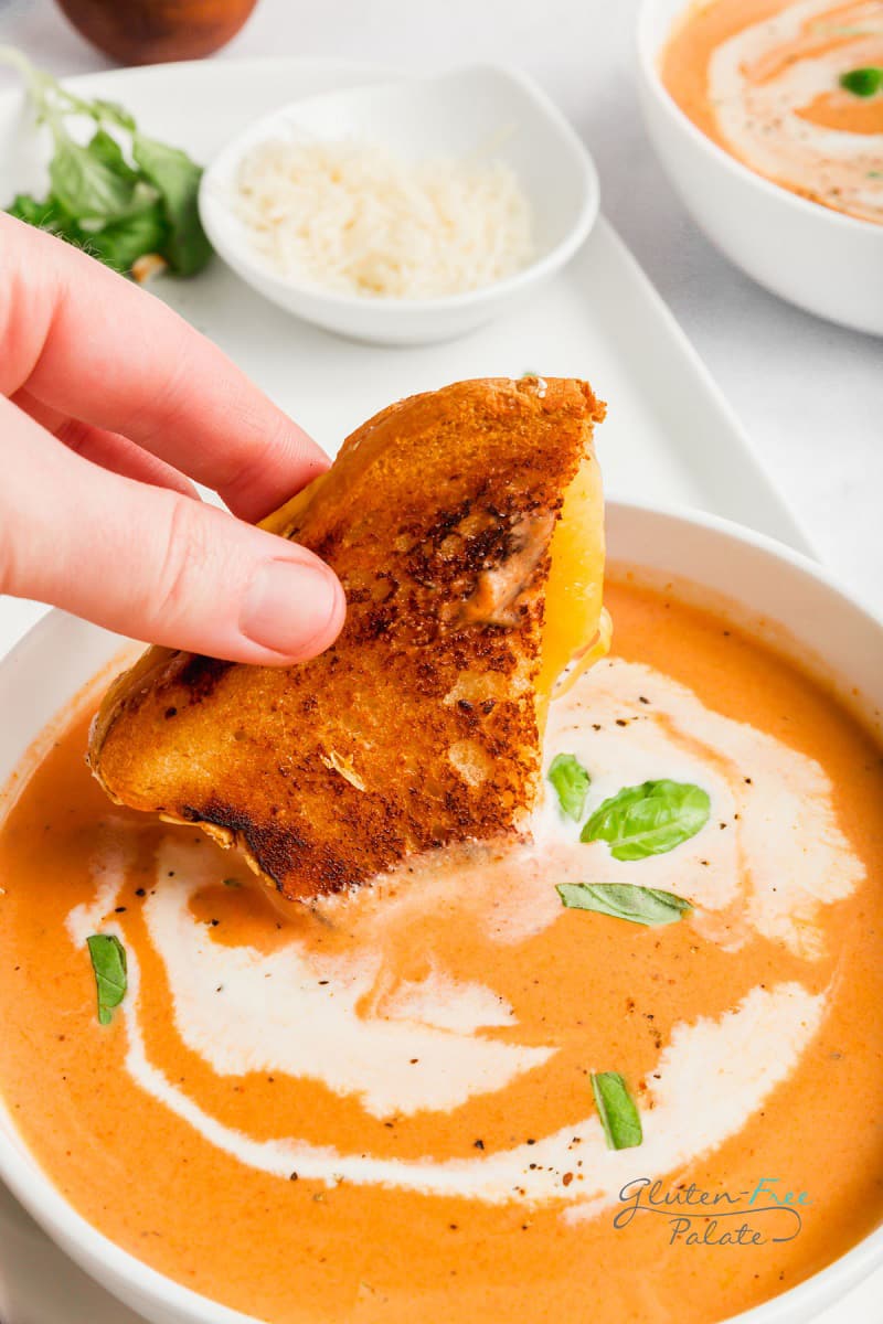 someone's hand dipping a grilled cheese sandwich into a bowl of creamy tomato soup.