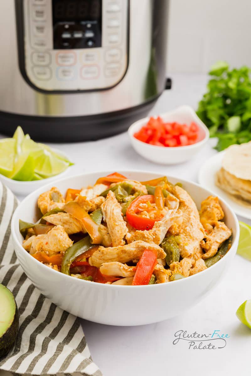 a large white bowl of sliced chicken, and red and green peppers in front of an instant pot and taco fillings.