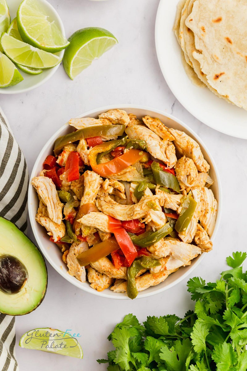 Top down view of a large bowl filled with chicken fajita filling, surrounded by tortillas, limes, avocado and cilantro