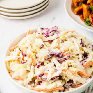 a bowl of creamy coleslaw on a table with a stack of plates and a dish of chicken.