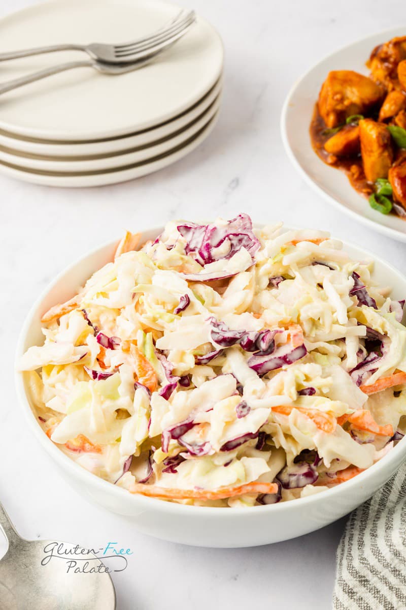 a bowl of creamy coleslaw on a table with a stack of plates and a dish of chicken.