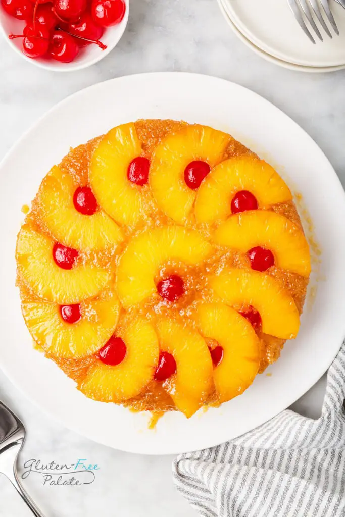 top down view of a round gluten-free pineapple upside down cake topped with pineapple ring halves and cherries on a white plate.