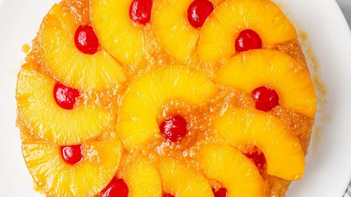 top down view of a round pineapple upside down cake topped with pineapple ring halves and cherries on a white plate.