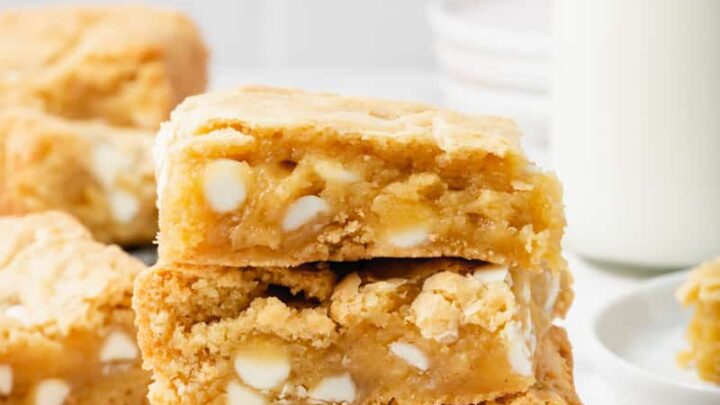 ThreeGluten-Free Blondies filled with white chocolate chips and stacked on top of each other. In the background is a glass bottle of milk and more blondie squares.