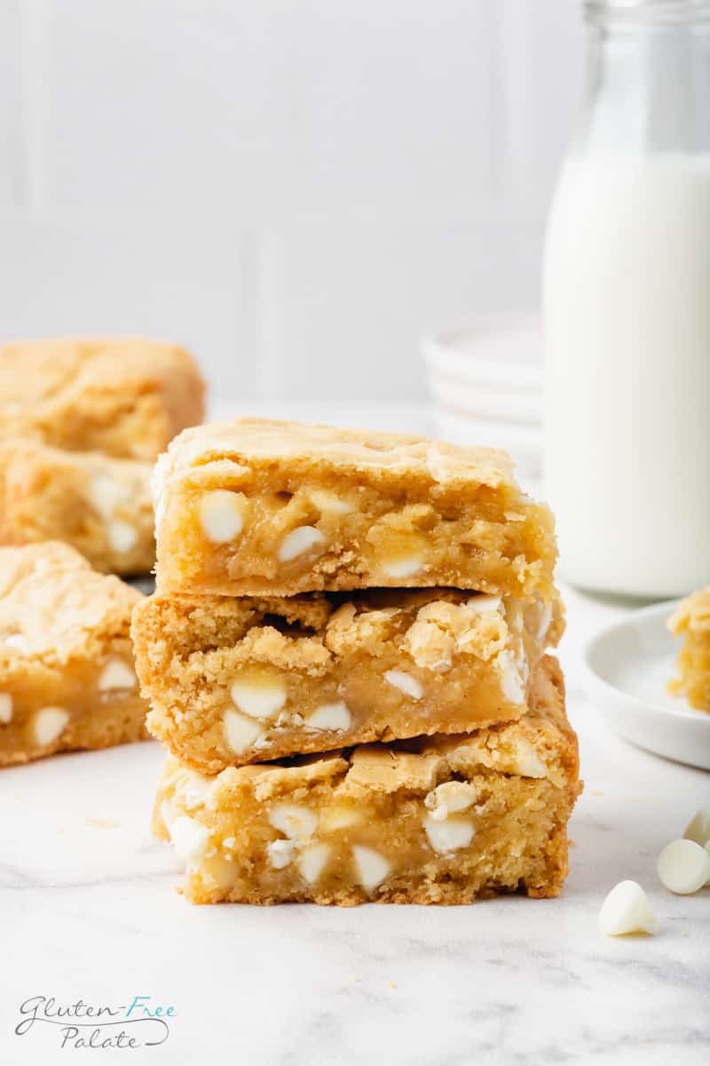 Three blondie bars filled with white chocolate chips and stacked on top of each other. In the background is a glass bottle of milk and more blondie squares.