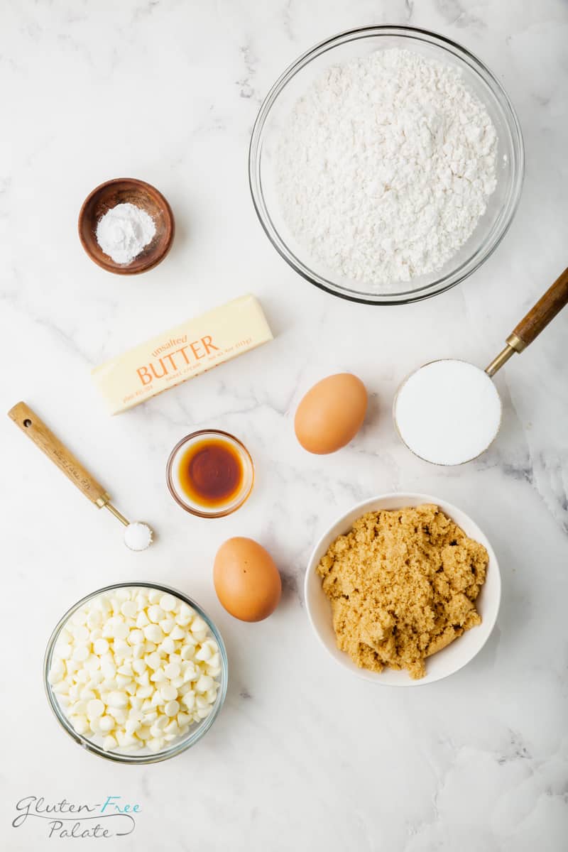 top down view of the ingredients needed to make Gluten-Free Blondies. There are two eggs, one stick of butter, a bowl of flour, a bowl of brown sugar, a bowl of white chocolate chips, a small bowl of vanilla extract and a small bowl of baking powder.