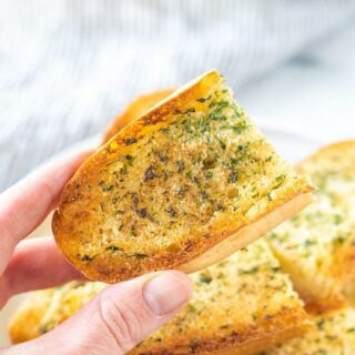 A hand holding an end piece of garlic bread over a plate filled with gluten-free garlic bread.