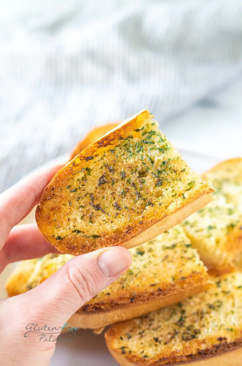 A hand holding an end piece of garlic bread over a plate filled with garlic bread.