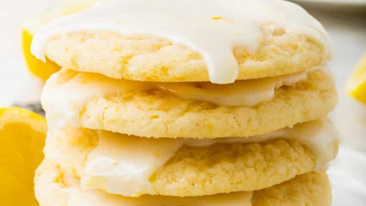 a stack of five gluten-free glazed lemon cookies stacked on a white plate. A lemon wedge is in front of the plate, and another lemon wedge is behind the stack of cookies.