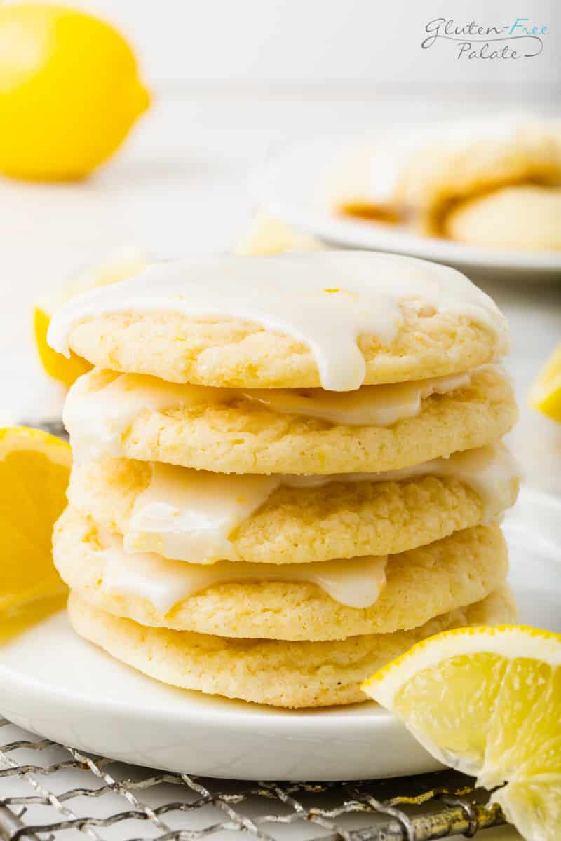 A stack of five glazed gluten-free lemon cookies stacked on a white plate. A lemon wedge is in front of the plate, and another lemon wedge is behind the stack of cookies.