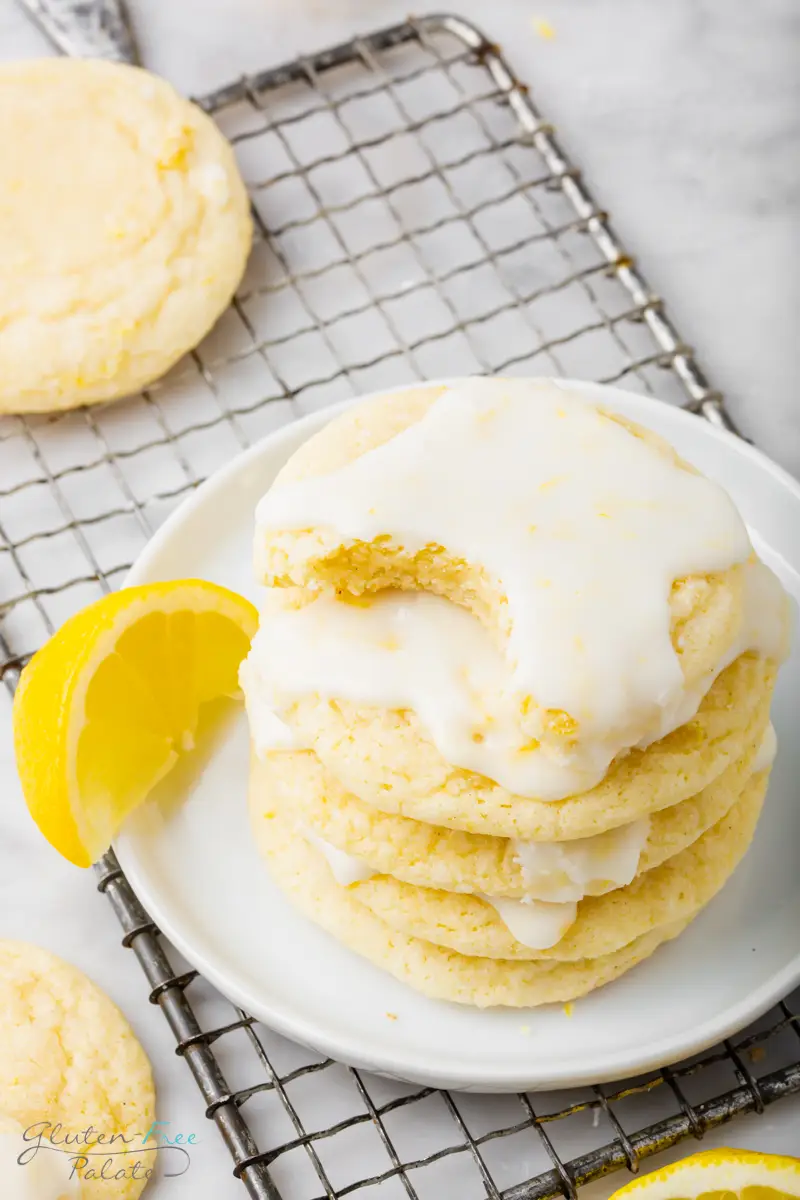 a white plate with five glazed lemon cookies n it. The top cookie has a bite missing. The plate is on top of a cooling rack and garnished with a lemon slice.